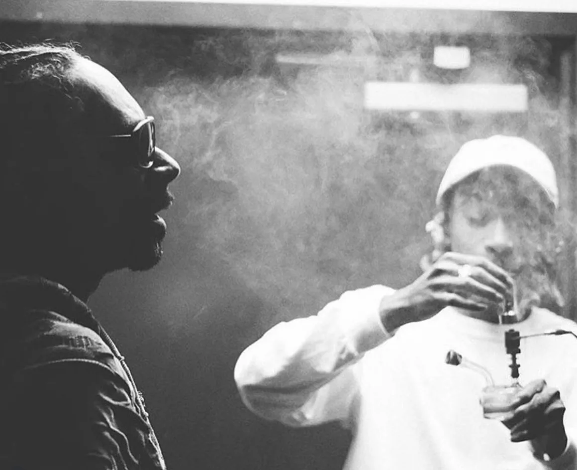 Snoop Dogg exhaling in the foreground while Wiz Khalifa takes a dab from an e-nail rig with a carb cap